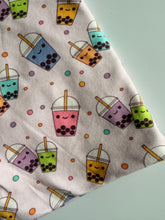 Load image into Gallery viewer, Close-up of a fabric item with an all-over novelty print featuring kawaii-style smiling bubble tea cups in pink, green, blue, purple, and orange with colorful polka dots scattered throughout, on a light cream
